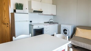 Eine kompakte Küche mit Kühlschrank, Herd, Spüle und Waschmaschine. Ein kleiner Esstisch und ein Bett mit Kissen sind vorhanden.