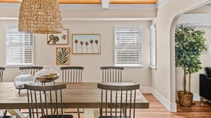 A dining area with a wooden table, chairs, and a woven pendant light. There are framed artworks on the wall and a potted plant in the corner.
