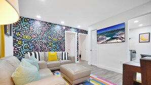 A modern living room with a colorful abstract wall mural, a large window with a cityscape view, and a striped rug. - 4 to 14 Guests 2 Kitchens Deco Retreat BBQ Patio (Miami)