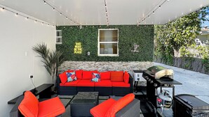 An outdoor patio with a red sofa, wicker chairs, a grill, and a stone wall with greenery. - 4 to 14 Guests 2 Kitchens Deco Retreat BBQ Patio (Miami)
