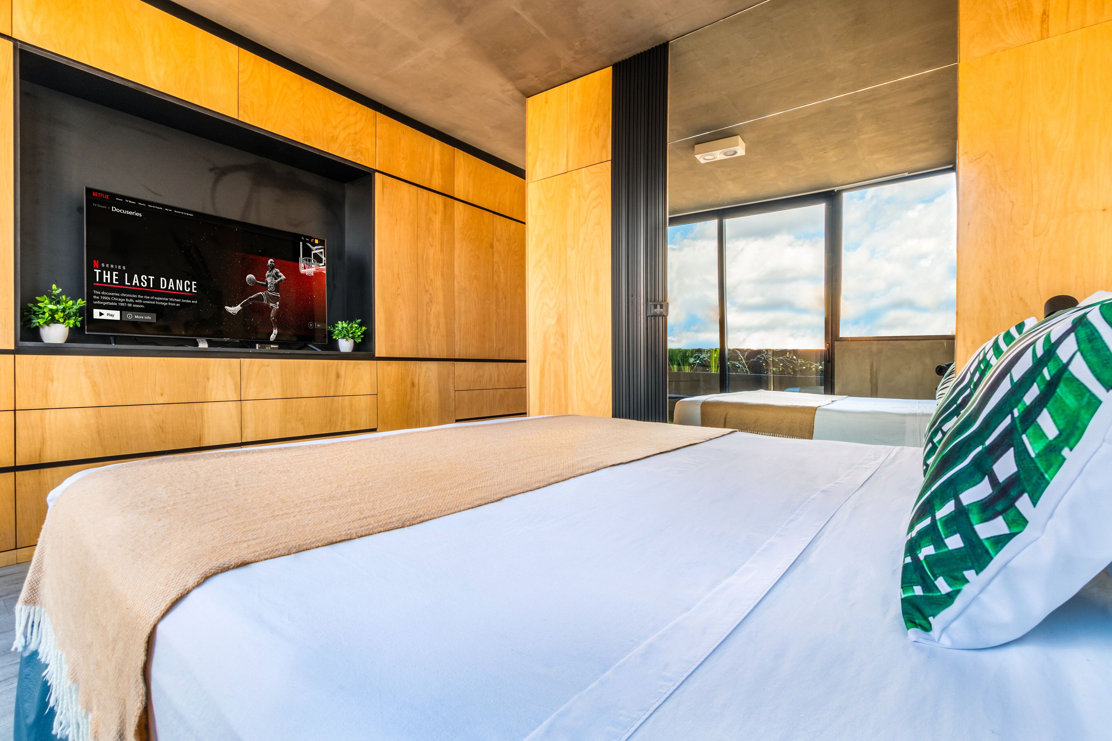 A modern bedroom with a large bed, a mounted TV displaying 'The Last Dance', and a view of the sky through large windows.