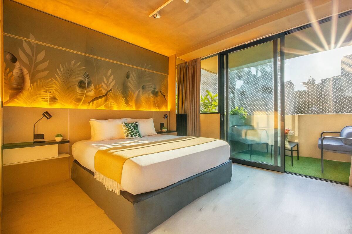 A modern bedroom with a large bed, a nightstand, a desk, and a view of the outdoors through a sliding glass door.