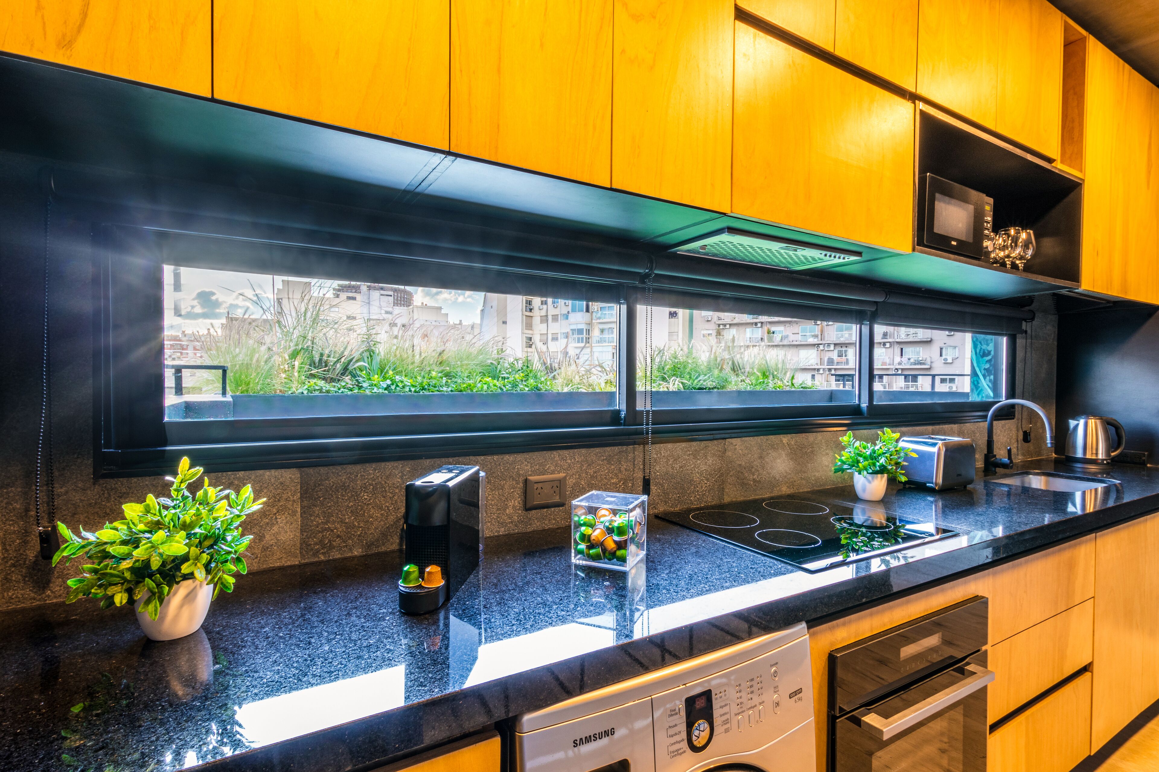 A modern kitchen with wooden cabinets, a black countertop, a built-in oven, a dishwasher, and a window with a view of buildings and greenery.