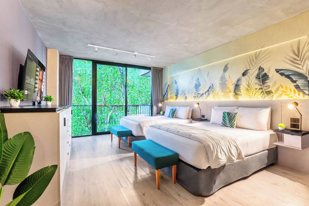 A modern bedroom with a large bed, two bedside tables, a bench, and a view of greenery through large windows.