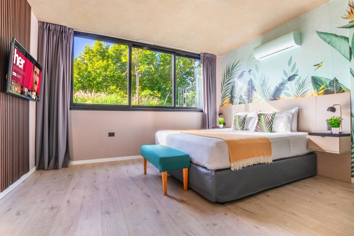 A modern bedroom with a large bed, a bench, a TV, and a window with a view of trees.