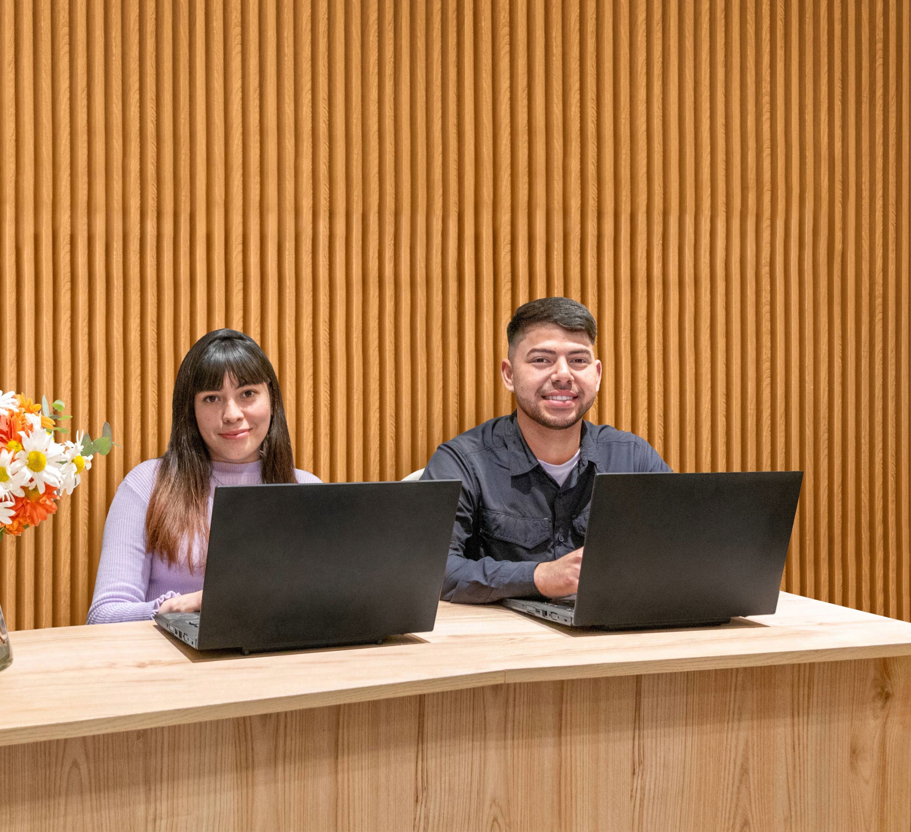 A reception desk with two people working on laptops, a vase of flowers, and a sign indicating the desk is open 24/7.