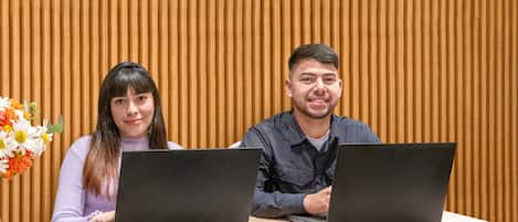 A reception desk with two people working on laptops, a vase of flowers, and a sign indicating the desk is open 24/7.