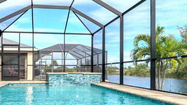 A swimming pool with a glass enclosure, a covered patio area, and a view of a body of water with palm trees. - Villa Teal | Heated Pool & Spa Coral Oaks Golf Course (Cape Coral)