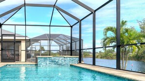 A swimming pool with a glass enclosure, a covered patio area, and a view of a body of water with palm trees. - Villa Teal | Heated Pool & Spa Coral Oaks Golf Course (Cape Coral)