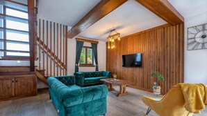 Un salon spacieux avec un canapé vert, un fauteuil jaune, un escalier en bois et un téléviseur fixé au mur.