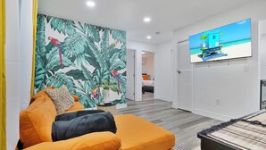 Ett modernt vardagsrum med en livfullt orange soffa, en stor väggmålning med tropiska blad och en platt-TV som visar en strandscen.