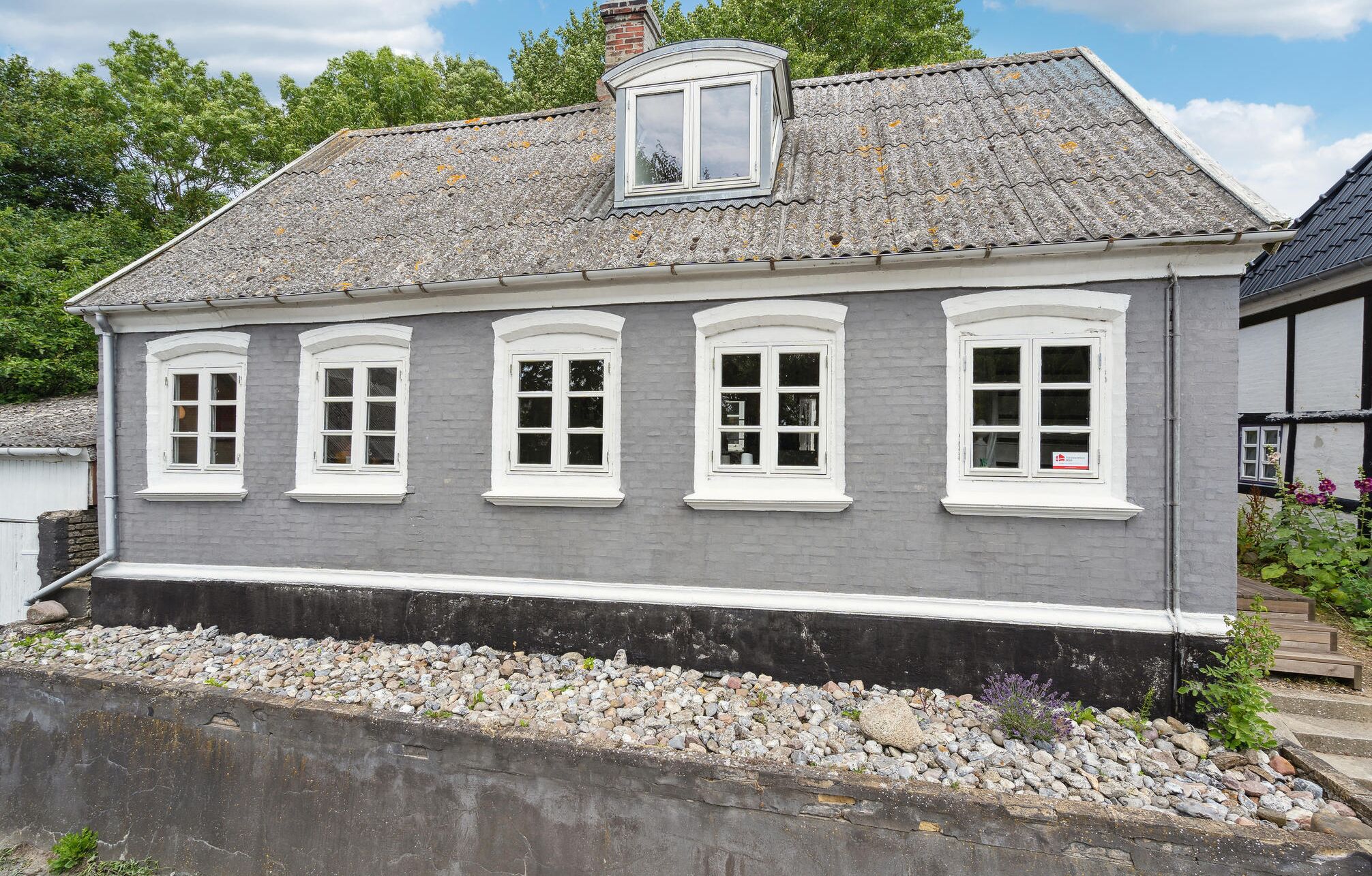 A two-story house with a grey facade and white trim, featuring five windows with white frames and a small dormer window above the central window.