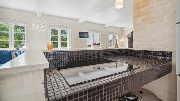 A modern kitchen with a black tiled backsplash, white countertops, and a built-in stove. There are windows with views of greenery outside, and a hanging light fixture above the stove.