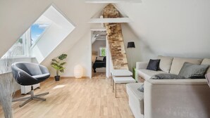 A modern loft bedroom with a sloped ceiling, a large skylight, a stone accent wall, a white sofa, a black chair, and a potted plant.