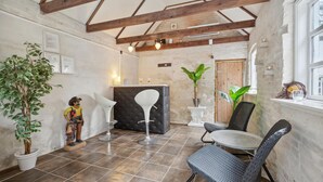 A rustic interior with exposed wooden beams, a tiled floor, a round table with chairs, and potted plants.