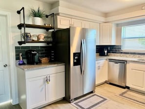 A modern kitchen with white cabinets, a stainless steel refrigerator, a coffee maker, and a window with blinds.