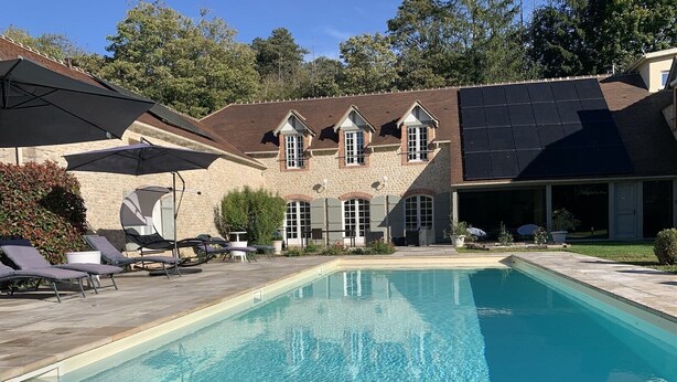 Una piscina con tumbonas y sombrillas, un edificio de piedra con garaje y paneles solares en el tejado.