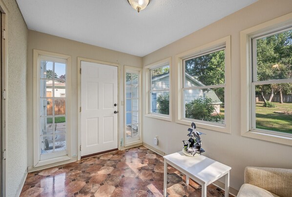 Una habitación soleada con suelo de baldosas, una puerta blanca y ventanas amplias que dan a un jardín.