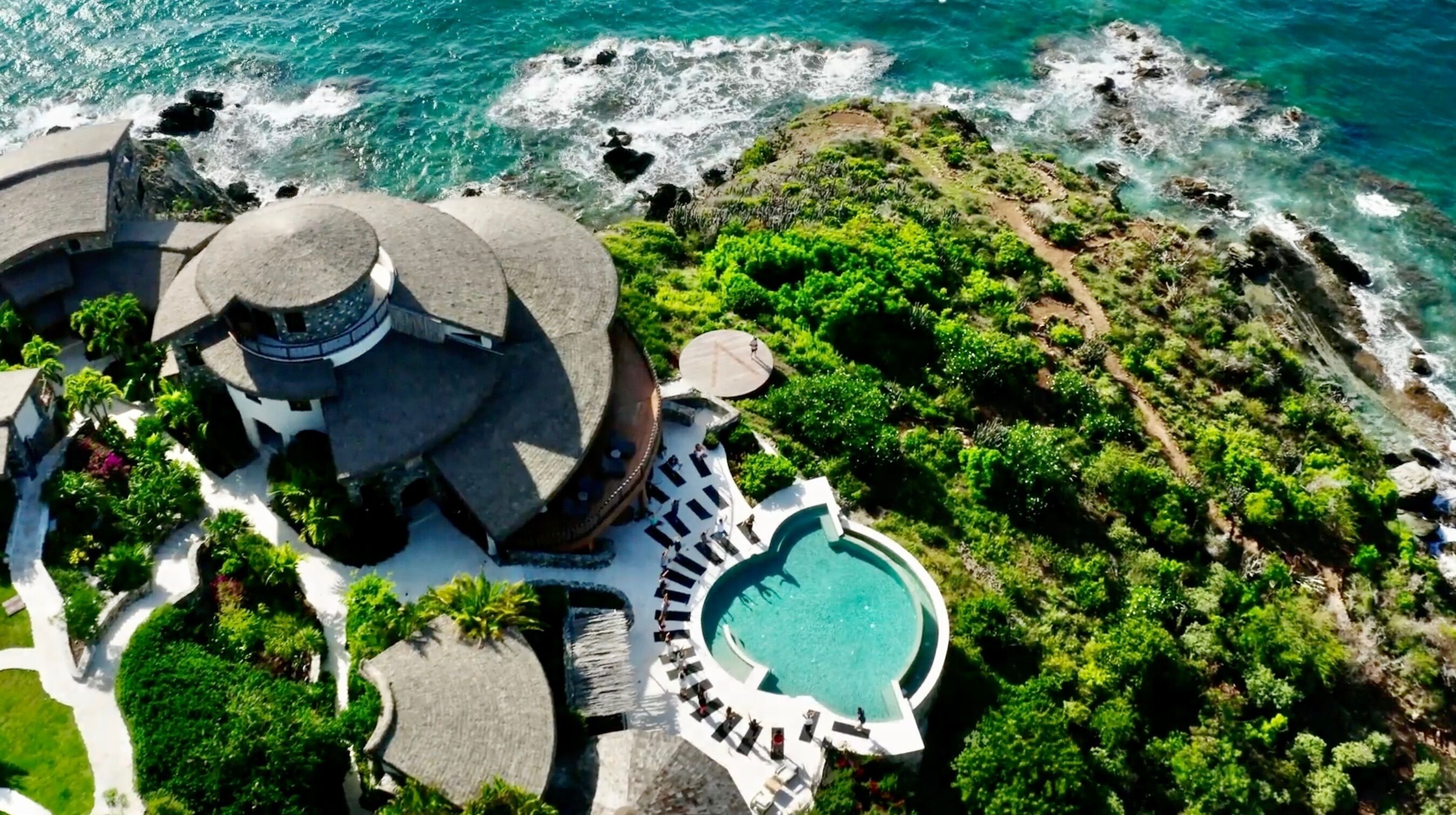 An aerial view of a resort with a pool surrounded by lounge chairs, a circular building, and a rocky coastline.