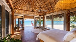 A room with a large bed, wooden flooring, a thatched ceiling, and a view of the beach. - The Aerial, BVI (East End)