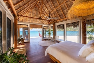 Una habitación con una cama grande, suelo de madera, techo de paja y vista a la playa.
