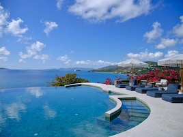 Una piscina infinita con vistas al mar y a las montañas, mobiliario exterior con sombrillas y un cielo azul despejado.