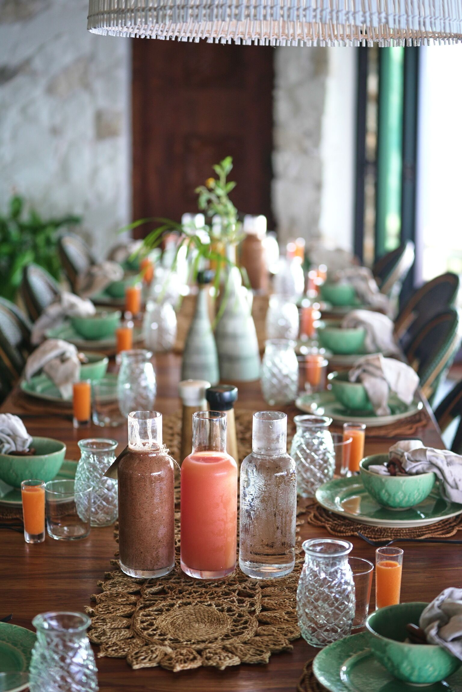 A long dining table set with green bowls, glasses, and bottles of beverages, with a woven placemat in the center.