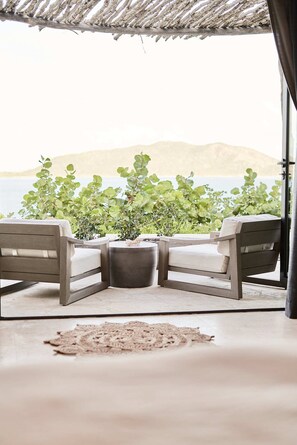 A corner seating area with two chairs and a table, surrounded by potted plants, overlooking a body of water and hills.