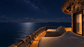 A starlit night view from a balcony with a thatched roof, wooden railing, and a cushioned seating area.