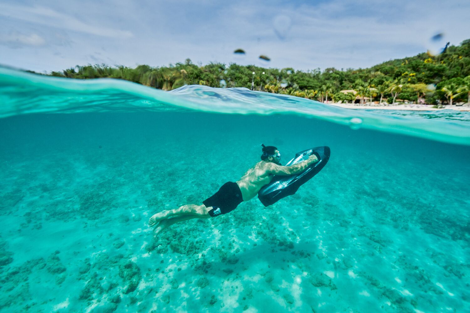 A person snorkeling underwater with a board, clear turquoise water, and a beach with palm trees and a hut in the background.