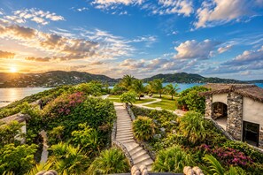 Un resort tropical con un sendero de piedra que conduce a una estructura de techo de paja, rodeado de exuberante follaje y con vistas a un paisaje costero.