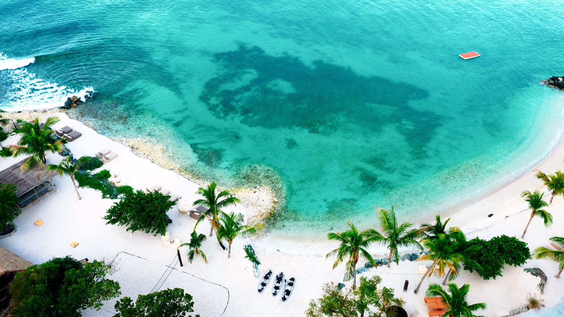 An aerial view of a beach with clear turquoise waters, white sand, and palm trees.