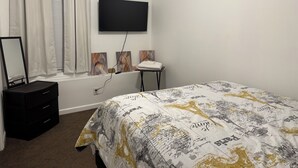 Un dormitorio con cama, cómoda, ventana con cortinas, televisor de pantalla plana y cuadros en la pared.