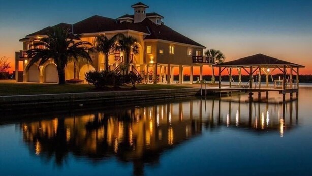 Una propiedad frente al agua con una casa grande, un muelle techado y una superficie acuática tranquila que refleja la edificación.