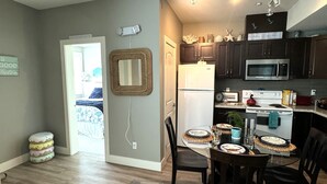 A modern kitchen with dark cabinets, a white refrigerator, and a microwave. There is a dining area with a table set for two, and a black leather couch in the foreground. - White Rock Beach Getaway (White Rock)