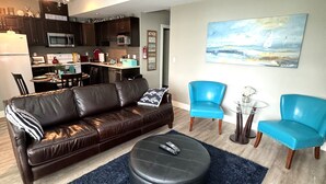 A living room with a brown leather sofa, two blue chairs, a black ottoman, and a painting of a beach scene. - White Rock Beach Getaway (White Rock)