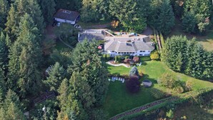 An aerial view of a large, multi-story house surrounded by dense forest, with a well-maintained lawn and a driveway leading to the entrance.