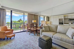Relax in a spacious living area with a sofa, ottoman, and dining set, offering a view of lush greenery and the sky.