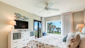 A bedroom with a bed, a TV, a ceiling fan, a dresser, and a view of palm trees. - Oceanfront Turtle Dunes Villa with Gorgeous Views! 2-Minute Walk to Sand! (Fernandina Beach)