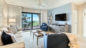 A living room with a sofa, armchair, coffee table, and a TV. There's a balcony with a view of palm trees and a pool. - Oceanfront Turtle Dunes Villa with Gorgeous Views! 2-Minute Walk to Sand! (Fernandina Beach)