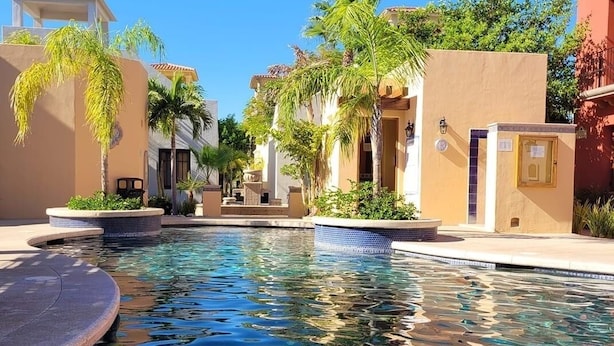 A pool area with a clear blue pool, surrounded by buildings with beige and white walls, palm trees, and a clear sky. - BeautifulHome, in front of the community pool By Casago (Loreto)