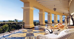 A covered outdoor area with a tiled floor, white wicker furniture, and a view of the ocean.