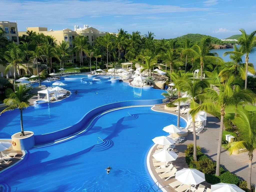 A resort pool with multiple levels and slides, surrounded by palm trees and lounge chairs.
