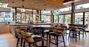 A bar area with a central round table surrounded by bar stools, a high ceiling with exposed beams, and a view of the outdoors through large windows.