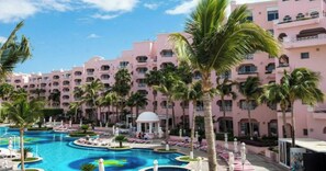 A resort with a large swimming pool surrounded by palm trees and a pink building with balconies.