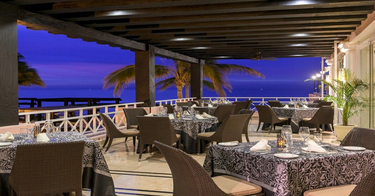 An outdoor dining area with tables and chairs, overlooking a body of water at dusk.