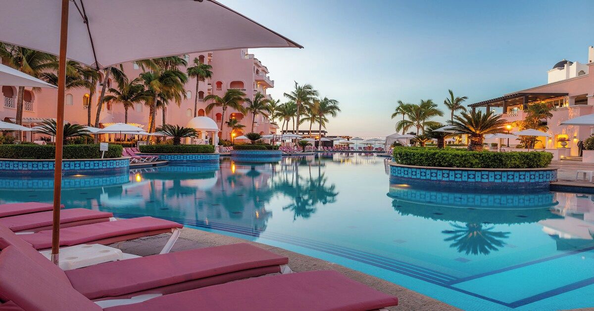 Zona de piscina con sillones de descanso y sombrillas, rodeada de palmeras y un edificio de fachada rosa claro.
