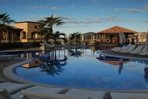 A swimming pool with lounge chairs and palm trees, surrounded by buildings with Mediterranean architecture.