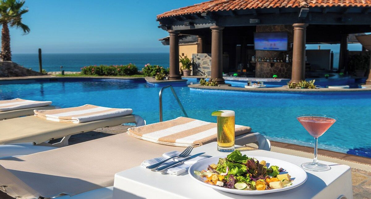 Un área de comedor junto a la piscina con vistas al océano, un bar y sillones de descanso.