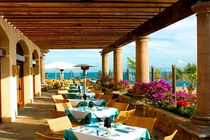 An outdoor dining area with tables and chairs, a wooden pergola, and a view of the sea.